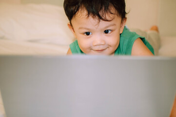 A male toddler is playing with a laptop while on the bed