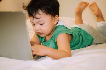 A male toddler is playing with a laptop while on the bed