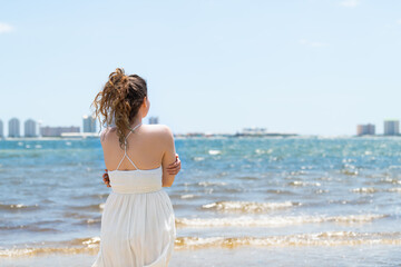 Young woman back cold shivering standing in white dress on beach bay shore in Florida with...