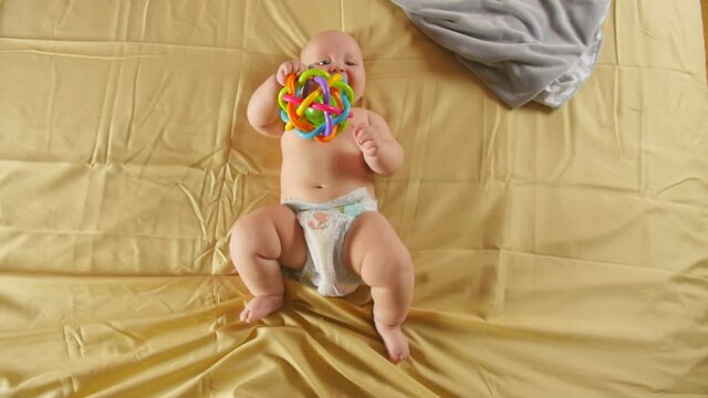Smiling baby lies on the bed, holds the toy with his hands and nibble a toy. Sensory development of a child. Full shot.