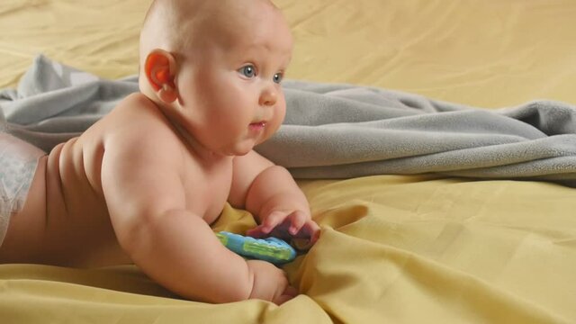 A chubby four months baby lies on his tummy and nibbles a toy. Sensory development of a child. Medium shot.