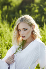 Girl in a long white dress sits on the grass in a field. Blonde woman in the sun in a light dress. Girl resting and dreaming, perfect summer makeup on her face