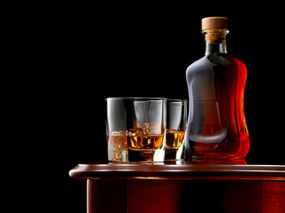 Two glasses of whiskey and ice on the wooden table. In the background, a full bottle of whiskey. Low key.