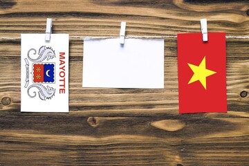 Hanging flags of Mayotte and Vietnam attached to rope with clothes pins with copy space on white note paper on wooden background.Diplomatic relations between countries.