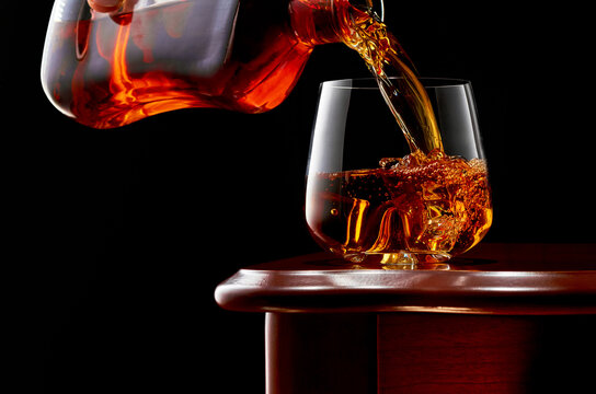 A Glass Of Whiskey With A Double Shot Of Whiskey And Ice On A Wooden Table. From Above In The Glass Is Poured Whisky From The Bottle. Close Up. Low Key.