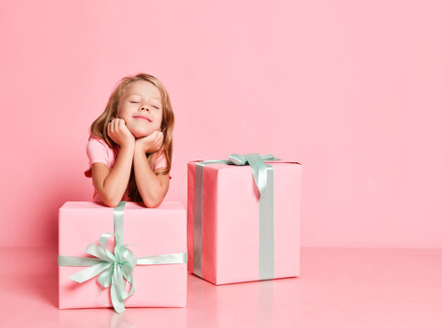 Christmas and New Year. Kid girl is sitting behind gifts with her chin in her hands, dreaming with closed eyes on pink