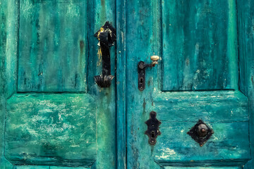 Close up of an old weathered and peeling blue painted external door with original worn wrought iron and brass furniture. With pitting, scratches and flaking. Lopud harbour, Croatia