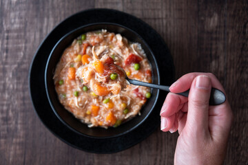 Woman’s hand with black metal spoon, Puerto Rican Chicken Rice Soup in a black stoneware bowl and saucer on a dark wood table