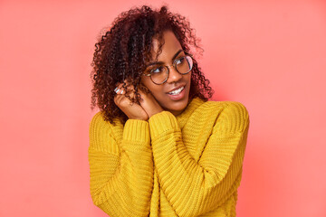 Happy pleased Afro American woman keeps hands together near face, focused away, notices desirable things, laughs, dressed in bright sweater, poses against pink background with free space aside