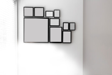 Group of various blank black picture frames on grey wall near a window with light, space for text