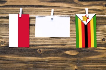 Hanging flags of Monaco and Zimbabwe attached to rope with clothes pins with copy space on white note paper on wooden background.Diplomatic relations between countries.