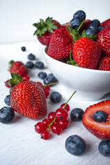 Fresh berries (strawberries, blueberries, red currant) in a white bowl on a white table, shallow depth of field