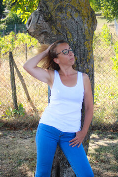 Young long-haired blonde woman leaning against a tree in summer in the sun. Sensual girl taking a lascivious pose in nature with hand in hair.