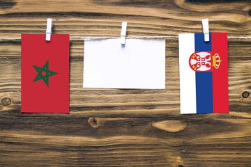 Hanging flags of Morocco and Serbia attached to rope with clothes pins with copy space on white note paper on wooden background.Diplomatic relations between countries.