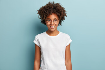 Waist up shot of happy curly woman with toothy smile, wears optical glasses and casual solid white...
