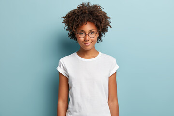 Attractive female student with curly hair, wears transparent glasses, white t shirt, stands against...