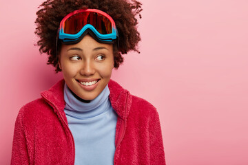 Pleased curly female traveler spends Christmas vacation in mountains, dressed in winter clothes, wears ski goggles on forehead, looks gladfully aside, models over pink wall. Active sport, leisure