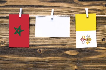 Hanging flags of Morocco and Vatican City attached to rope with clothes pins with copy space on white note paper on wooden background.Diplomatic relations between countries.