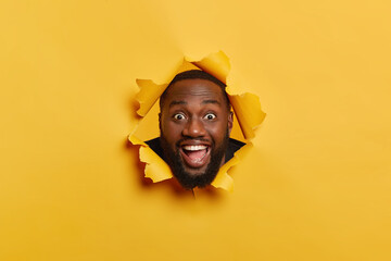 Photo of happy black man with pleased face expression, dark bristle, has fun indoor, keeps head in torn paper hole, laughs and looks at camera, isolated over yellow background. Face expression