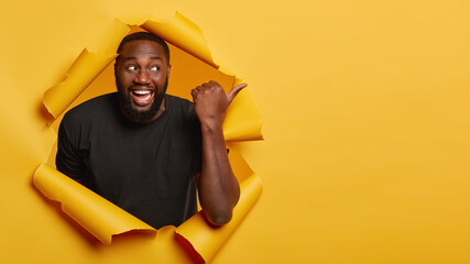 Cheerful handsome man with toothy broad smile, points aside, promots company product, gives advice to check out cool advertisement or awesome place, stands in paper torn hole, wears black clothing.