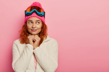 Winter sport concept. Attractive active woman has dreamy expression, recreats in mountains, thinks about snowboarding, wears pink hat with ski goggles, imagines ride on snowy slope, copy space