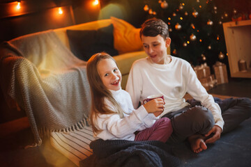 Obraz na płótnie Canvas Happy boy and girl friends or brother and sister with cups of cocoa with marshmallows in Christmas interiors with lights. The concept of Christmas and New Year.