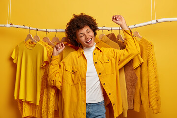Carefree curly woman clothes buyer dances with happiness, raises arms, buys yellow clothing from new collection, rejoicing successful shopping day, being in high spirit, dances against outfits on rack