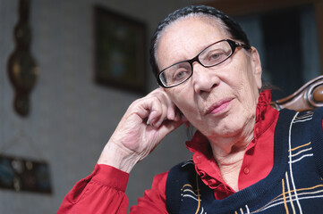 Portrait of an elderly pensive woman in glasses leaning on her arm indoors sitting and thinking. Elderly woman 80 years old