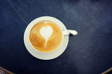 Heart foam latte coffee in white cup with spoon on black wooden round table background