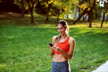 Attractive fit caucasian brunette in sportswear and with ponytail standing in nature, using smart phone and having earphones in ears. Sunny day in nature.