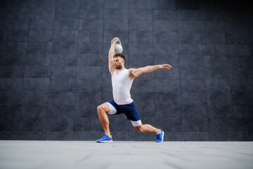 Fototapeta na wymiar Handsome strong muscular caucasian man in shorts and t-shirt doing lunges and holding kettle bell. In background is gray wall.