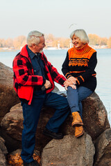 Cheerful elderly people a woman and a man are sitting on the stones and hugging on the lake,...