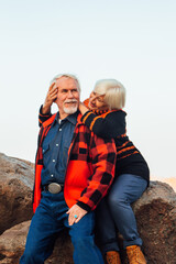 Fototapeta premium Cheerful elderly people a woman and a man are sitting on the stones and hugging on the lake, against the background of the bridge.