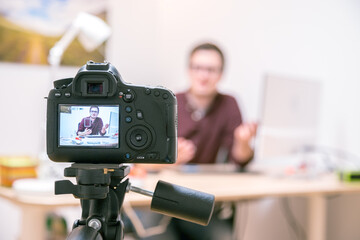 Video blogger at home: Camera on tripod is filming a young man sitting on his working place