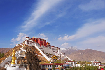 Lateral view of the Potala Palace in Lhasa, Tibet, surrounded by green vegetation, against a blue summer sky covered by white clouds.