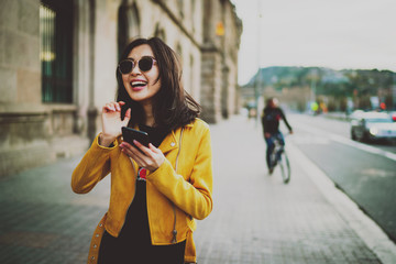 Half length portrait of beautiful laughing asian woman in a sunglasses with long dark hair wearing yellow leather jacket chatting online by a mobile phone while standing on blurred street background.