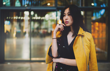 Fototapeta premium Attractive asian female with long dark hair looking pensively aside while standing on an urban background. Hipster girl waiting for the friends next to the shopping mall entrance to spend spare time