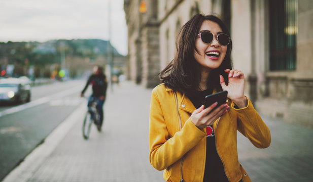 Cheerful asian student girl wearing modern sunglasses laughing at friends' photos in social media by a mobile phone. Happy model look woman in casual outfit checking blog comments via smartphone.