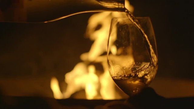 Hand pours white wine into glass in close up, foreground, in front of a fire pit burning in the background. Slow motion HD recorded at 180fps.
