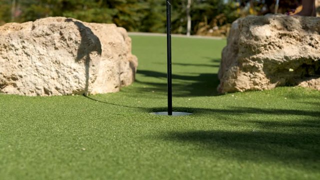 Blue ball passes near the mini golfing hole. This close up shot showing two rocks, mini golfing hole and stick. You can see the golf ball missing the hole and girl&rsquo;s feet with shoes passing nearby.
