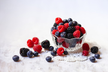 Ripe seasonal berries in glass bowl: raspberry, blueberry, blackberry. Healthy and no calories snack. White wooden background. Copy space for text