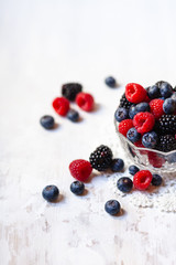 Ripe seasonal berries in glass bowl: raspberry, blueberry, blackberry. Healthy and no calories snack. White wooden background. Copy space for text