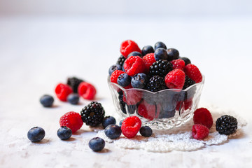 Ripe seasonal berries in glass bowl: raspberry, blueberry, blackberry. Healthy and no calories snack. White wooden background. Copy space for text