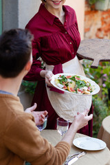 In the italian restaurant: young beautiful slim waitress in red dress is serving a salad to a man sitting at the table