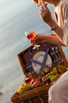 Young girl is drinking rose wine near the sea. Picnic basket with fruits in front. Relaxed atmosphere. Italy, Cinque Terre.