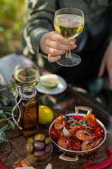 Young girl is holding a glass of white wine in her hand. Delicious italian food on the table: fried spicy shrimps in copper pan, olive oil, bread, olives. Light healthy meal, tasty snack