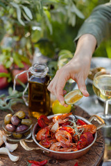 Girl is squeezing slice of lemon on delicious and spicy shrimps fried in copper pan with herbs, tomatoes and garlic. Wine, olives on the table. Luxury lifestyle, gourmet food for lunch