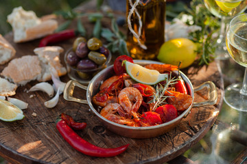 Romantic italian lunch outside for a couple: copper pan with delicious and spicy fried shrimps with herbs and garlic, wine, bread, olives. Bottle of olive oil. Luxury lifestyle, gourmet food