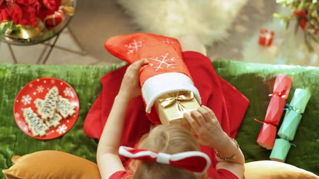 Holiday Season. Upper view of elegant little princess in red dress sitting on sofa in the modern living room at Christmas taking a Christmas present box out of a sock. PRORes 422 codec.