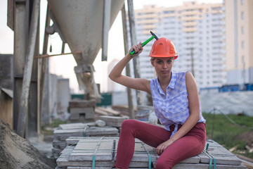 Cute young woman in construction helmet with hammer sits on cement and concrete slabs on construction site and hits yourself hammer on helmet